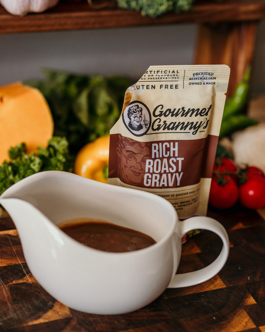 Gourmet Granny's Rich Roast Gravy packet with a white gravy boat filled with gravy on a wooden surface with vegetables in the background.